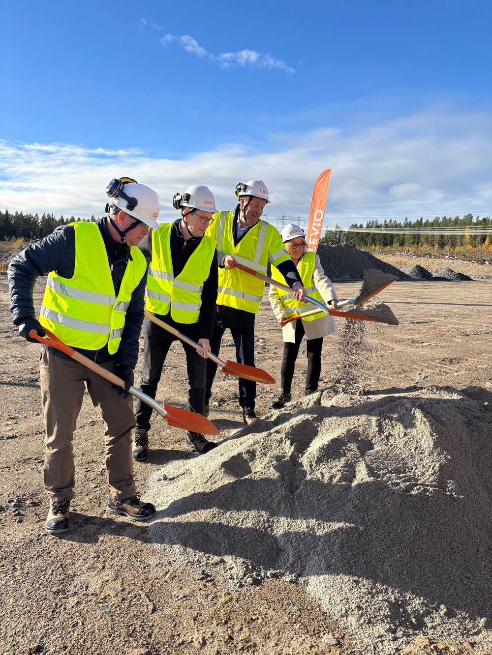 Fyra personer i bygghjälmar och reflexvästar tar första spadtaget med varsin spade på en byggarbetsplats.