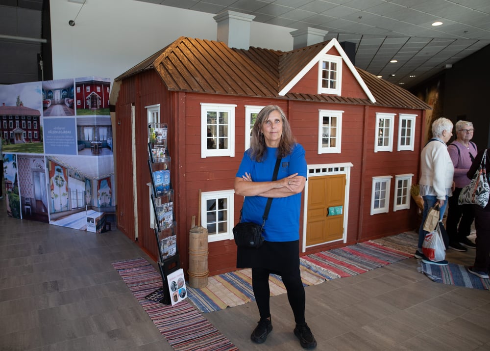Kvinna i blå tröja står framför en röd modell av en traditionell Hälsingegård.