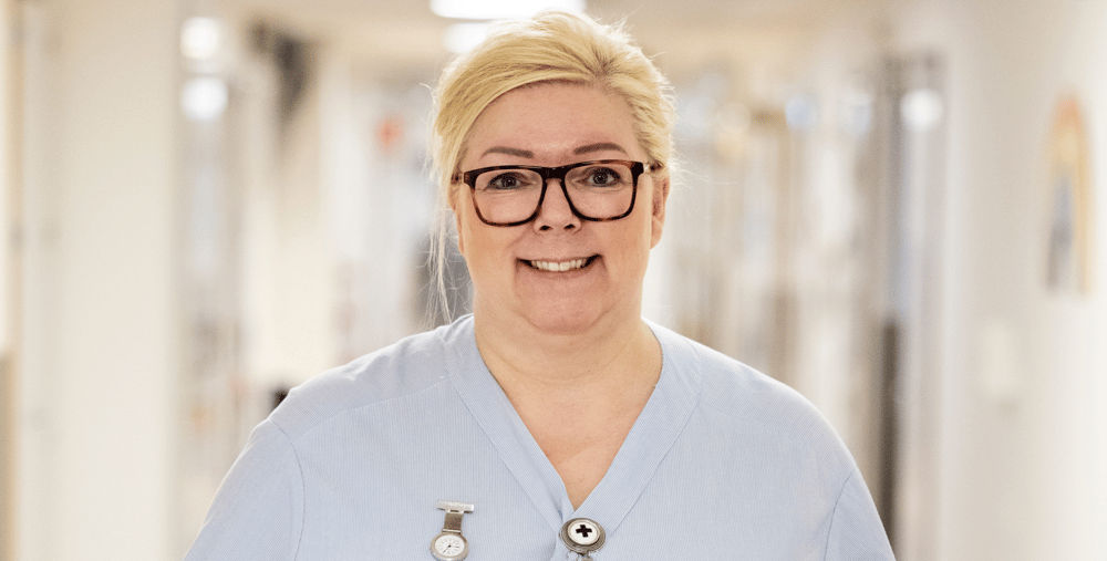 En kvinna i vårdkläder och glasögon står i en korridor.