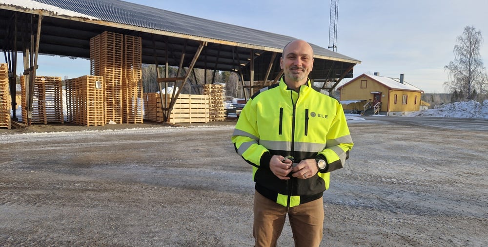 Mattias Ringström iklädd gul varseljacka står på en industriell gårdsplan med staplade träpallar och ett skjul i bakgrunden.