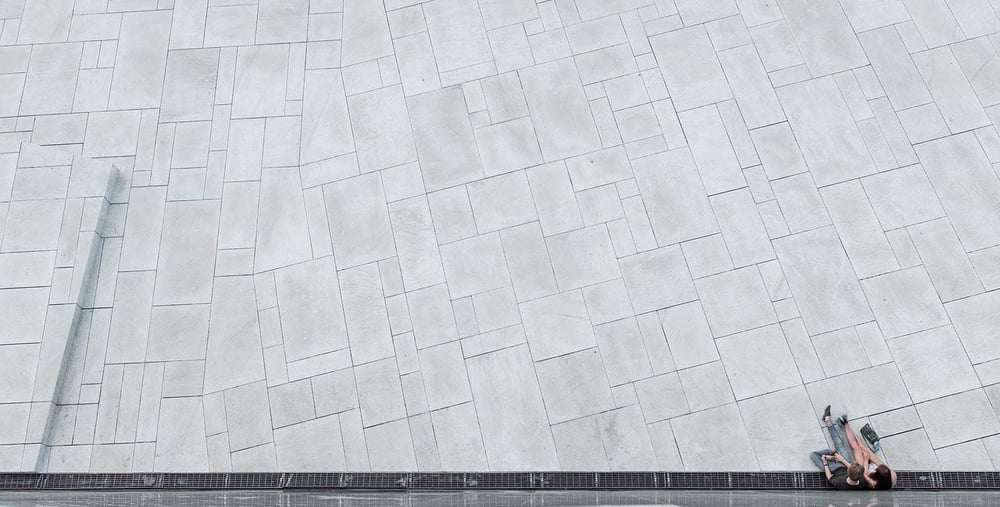 Två personer sitter på marken intill en speglande vägg på ett stort ljusgrått stenlagt torg.