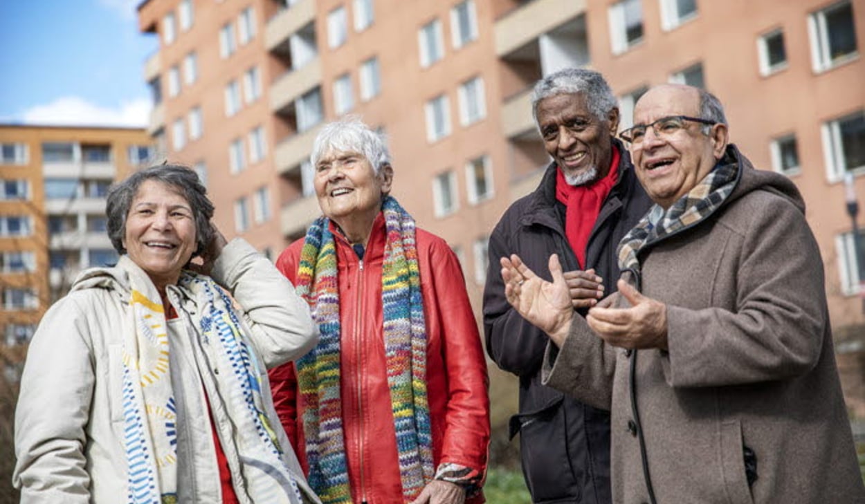 Fyra äldre personer står utomhus och samtalar framför ett bostadshus.