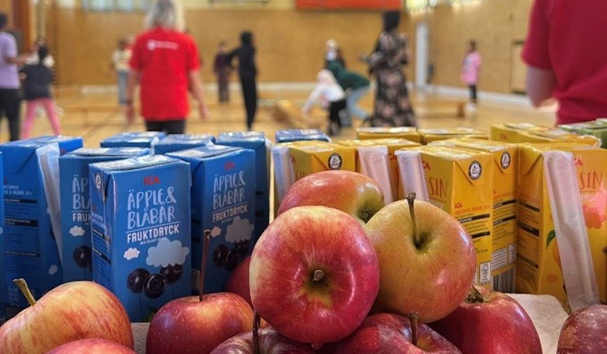 Äpplen och festisförpackningar i förgrunden, lekande barn i bakgrunden i en gymnastiksal