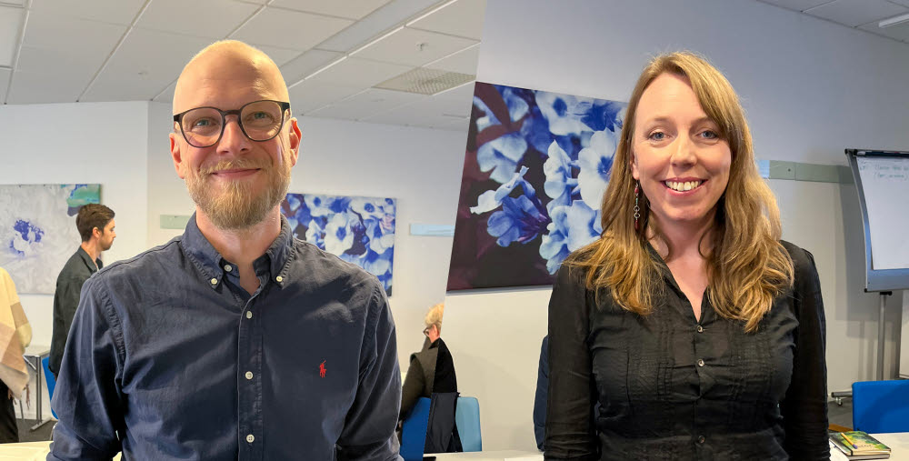 En man med glasögon, skägg och mörkblå skjorta står leende i förgrunden i ett konferensrum. Bredvid står en leende kvinna och i bakgrunden syns en målning med blå och vita blommor samt en blädderblockstavla.