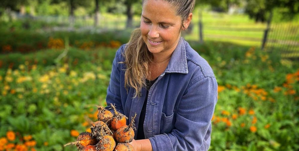 Person stående vid ett trädgårdsland och håller i grönsaker