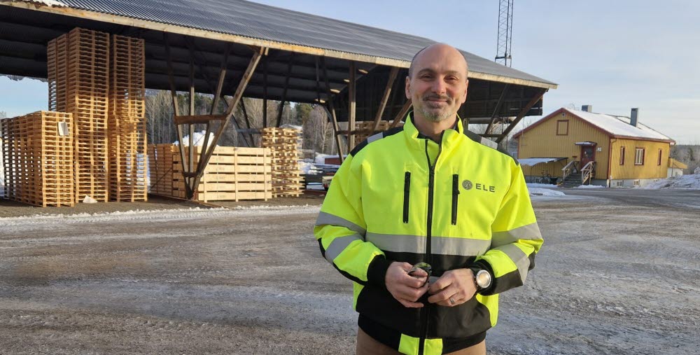 Mattias Ringström iklädd gul varseljacka står på en industriell gårdsplan med staplade träpallar och ett skjul i bakgrunden.