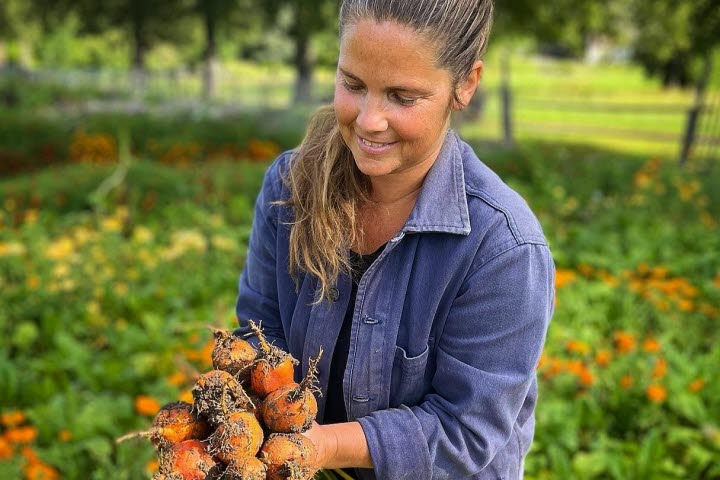 Person stående vid ett trädgårdsland och håller i grönsaker