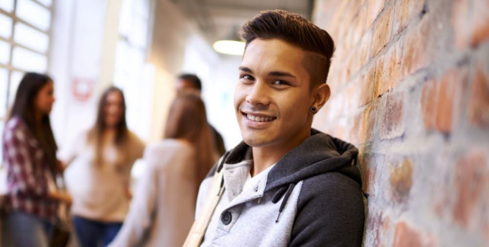 Ung person som st&#xE5;r mot en tegelv&#xE4;gg, fyra personer st&#xE5;r i bakgrunden och pratar med varandra. 