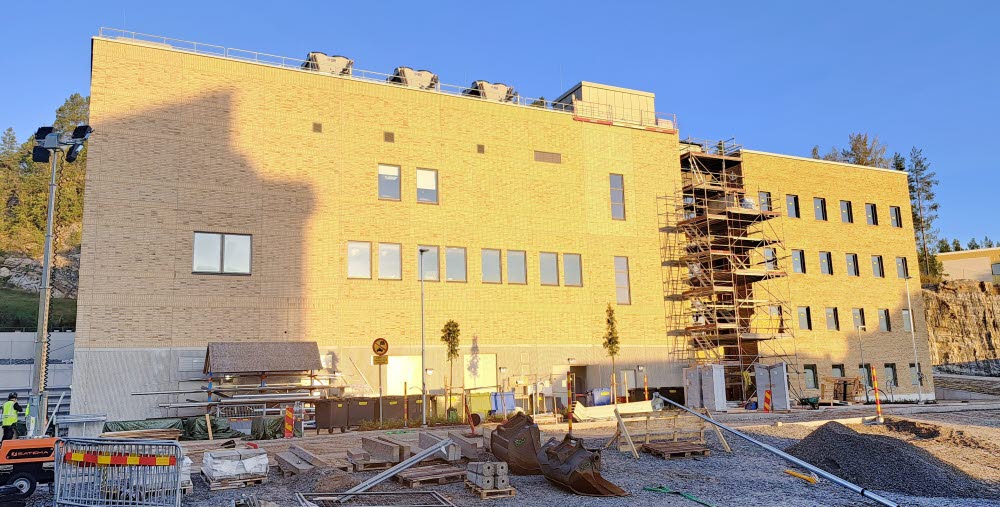 Bilden visar den nya teknikbyggnaden med administrativ byggnad på Hudiksvalls sjukhus. Himlen är blå och molnfri, i förgrunden finns byggarbetsmaterial. Byggnaden är en del av Framtidsbyggets projekt på Hudiksvalls sjukhus. 