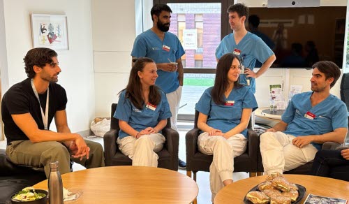 Samlingslokal med vita väggar, två bruna bord, sju personer som sitter och samtalar. Fyra personer i blåa och vita kläder och två personer i privata kläder. AT/BT centrum